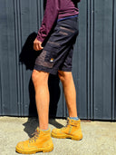 Person wearing black cargo work shorts and brown work boots against a gray wall.