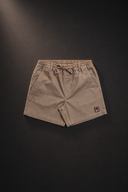 A pair of beige drawstring shorts with a small orange and black logo patch on the lower left hem, displayed flat against a dark background.