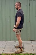 A man shown in profile standing outdoors in front of a green corrugated metal wall. He is wearing a dark charcoal t-shirt, khaki cargo shorts, and brown work boots.
