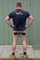 A man stands with his back to the camera wearing a black t-shirt with a white 'BT' logo on the back, khaki shorts, and boots, posing in front of a green corrugated metal wall.