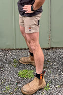A person wearing beige work shorts with a DFP LOGO on the left leg, paired with tan work boots and black socks, standing on a gravel surface in front of a green metal door.