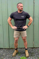 A man wearing a dark charcoal gray t-shirt with a white 'BT' logo on the chest, khaki shorts, and tan boots, standing in front of a green corrugated metal wall.